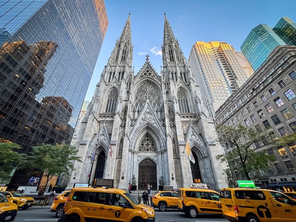 St. Patrick's Cathedral with yellow taxis parked outside, framed by high-rise buildings in New York City.