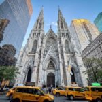 St. Patrick's Cathedral with yellow taxis parked outside, framed by high-rise buildings in New York City.