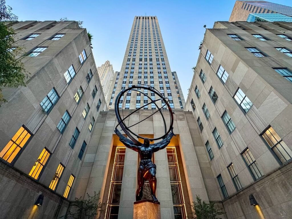 A low-angle shot of the Atlas bronze statue. The sculpture stands between tall skyscrapers against a blue sky.