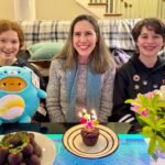 Three people smile together with a cupcake bearing lit candles and a colorful flower arrangement in the front.