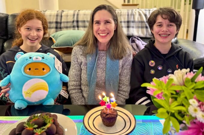 Three people smile together with a cupcake bearing lit candles and a colorful flower arrangement in the front.
