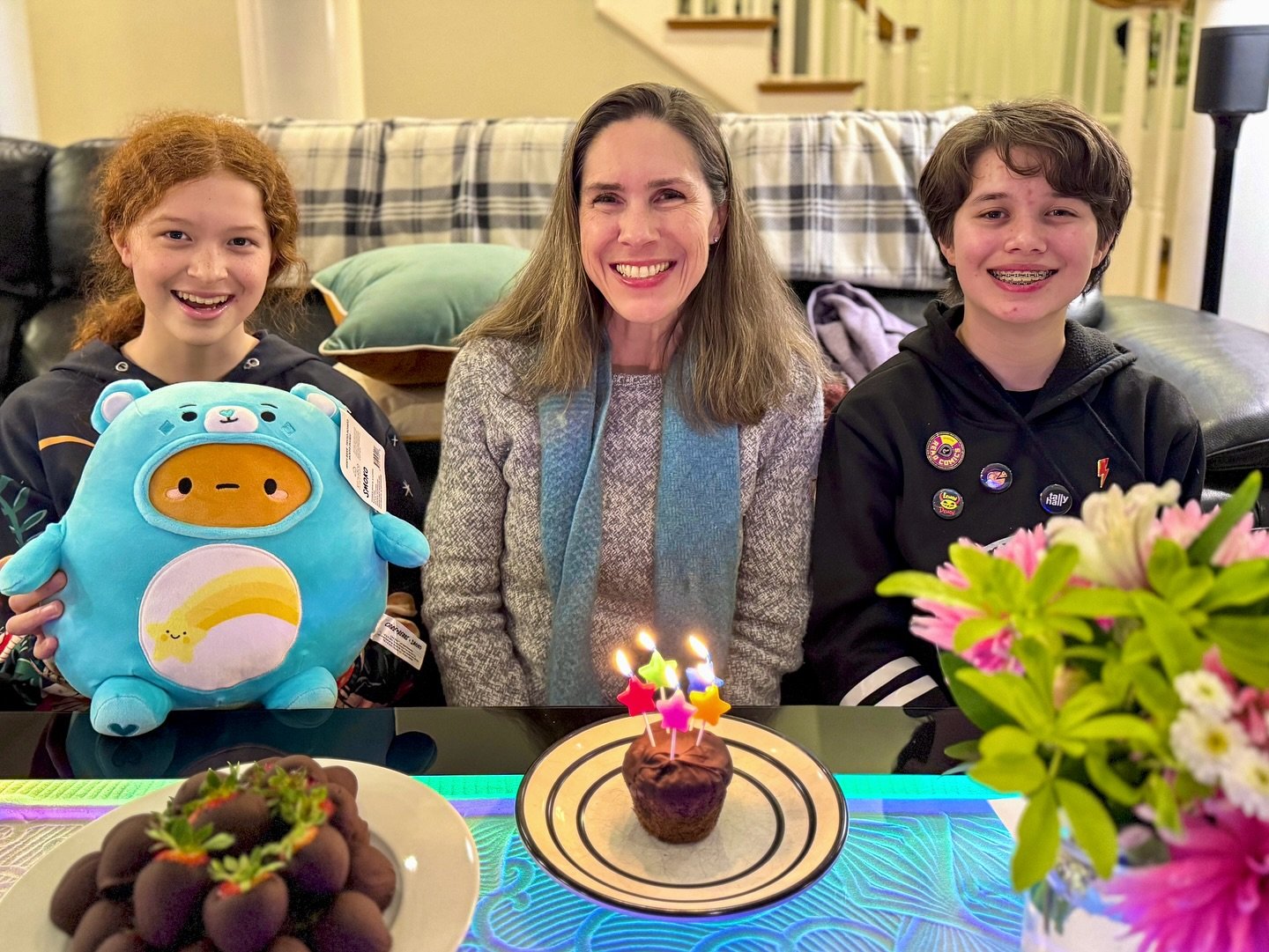 Happy Birthday Three people smile together with a cupcake bearing lit candles and a colorful flower arrangement in the front.