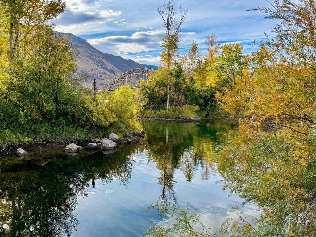 A serene river scene with vibrant autumn foliage reflected on the water. Rugged mountains and rocky banks frame the landscape under a partly cloudy sky.