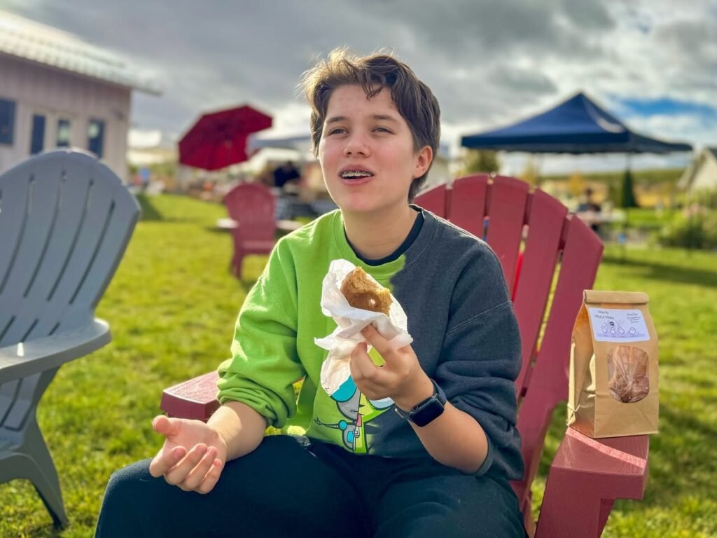 A person sitting in an Adirondack chair holding a wrapped pastry outdoors. In the background, a red umbrella and a blue tent are visible.