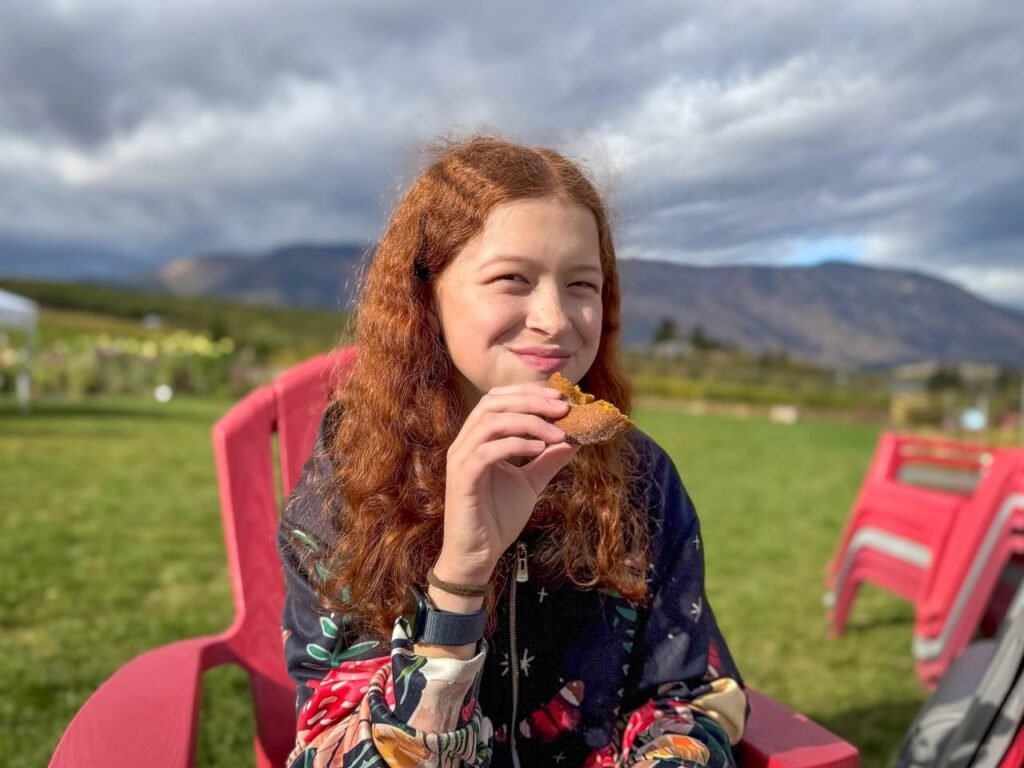 A person with red hair sits on a red chair eating a snack while enjoying a scenic outdoor view. The background features rolling hills and a dramatic, cloudy sky.