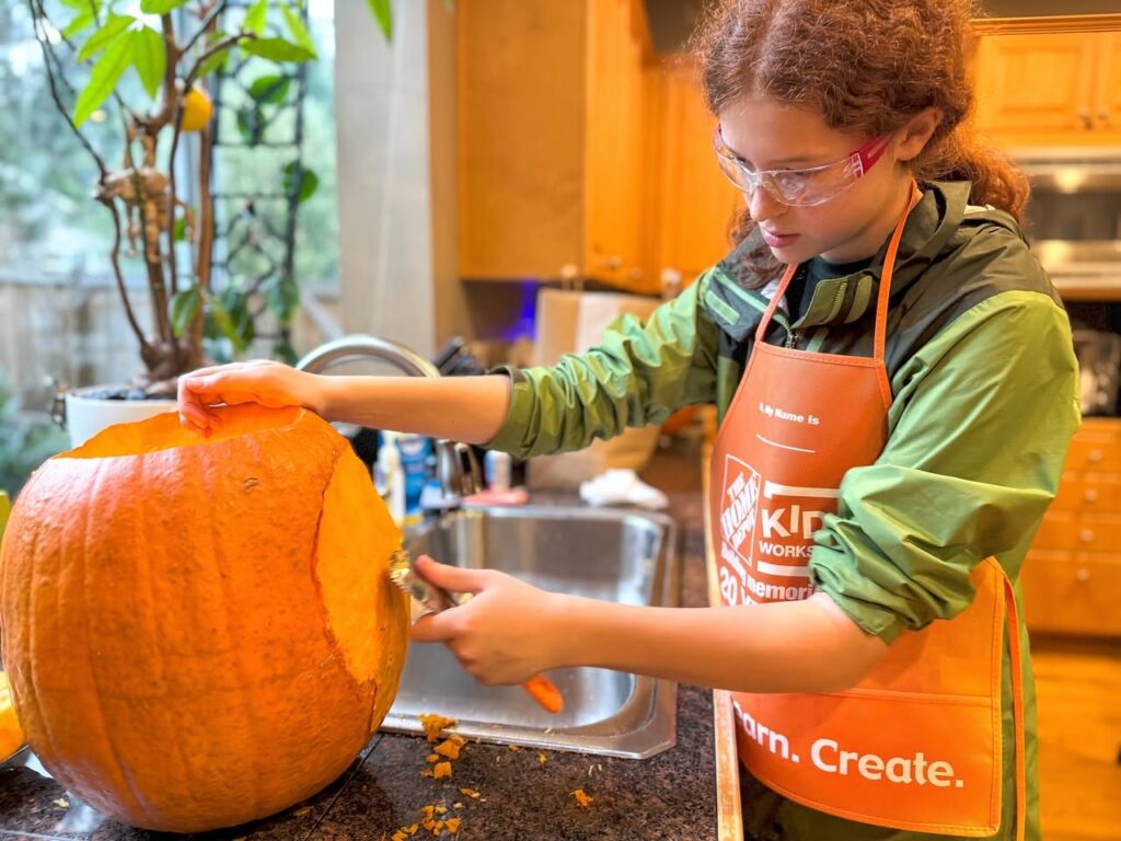 A person wearing safety glasses and an apron focuses on carving a large orange pumpkin in a cozy kitchen.