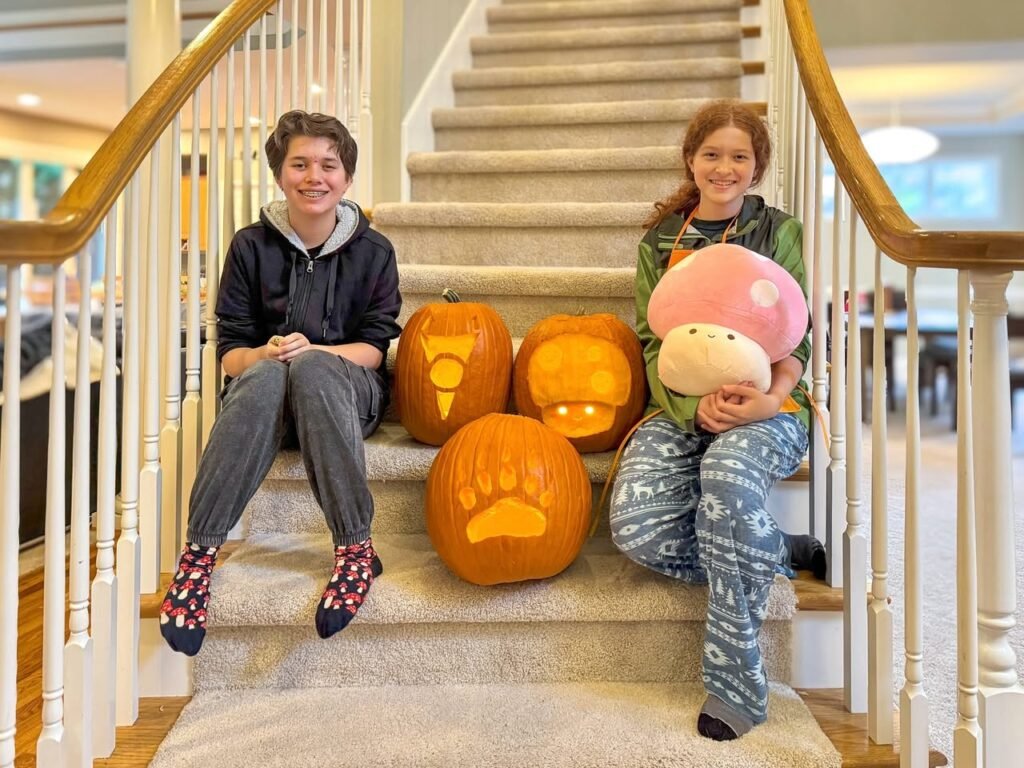 Two young people pose with glowing carved pumpkins on a staircase in a home.