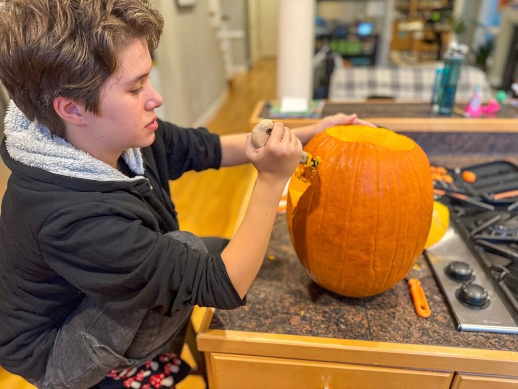 A person concentrates while carving a pumpkin on a kitchen counter. A detailed view shows the hand holding the tool near the orange gourd in a cozy home setting.