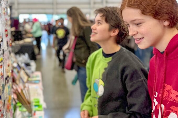 Two young people browsing handmade goods at a local market stall. The background shows a bustling community event with other visitors.