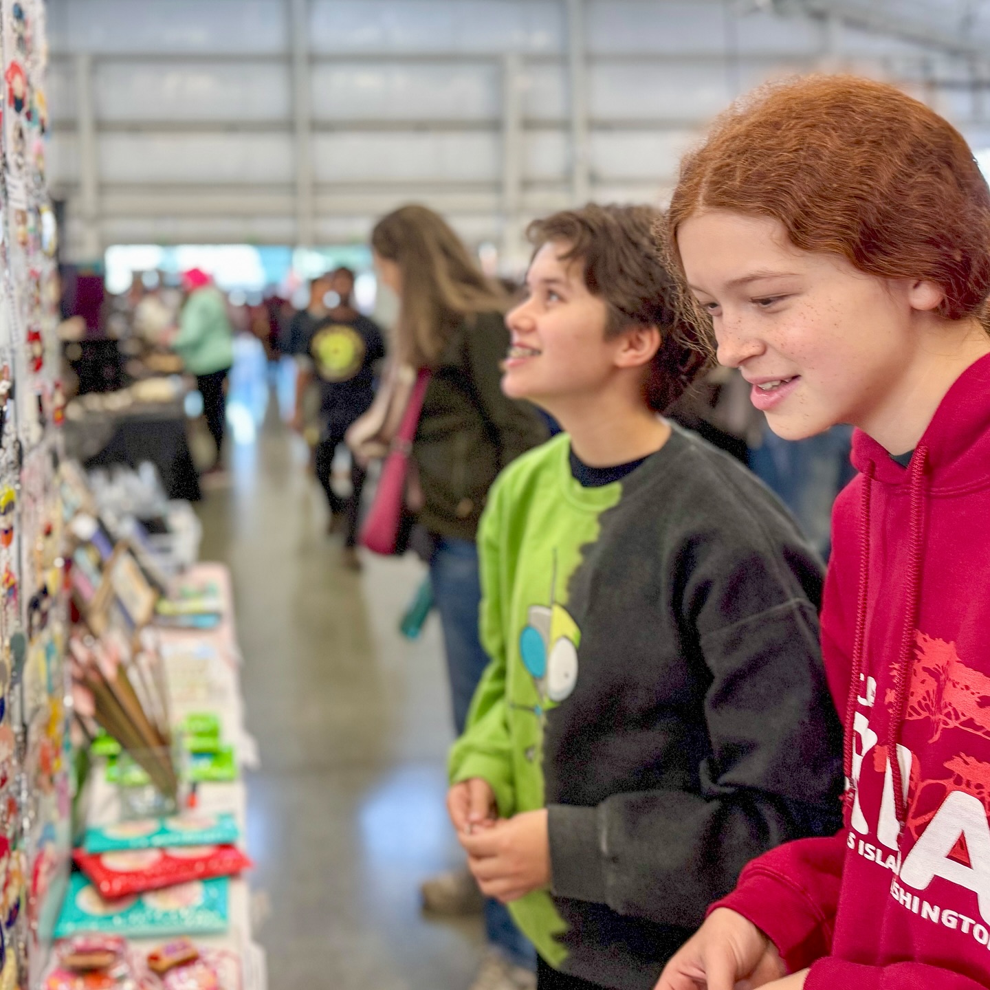 Two young people browsing handmade goods at a local market stall. The background shows a bustling community event with other visitors.
