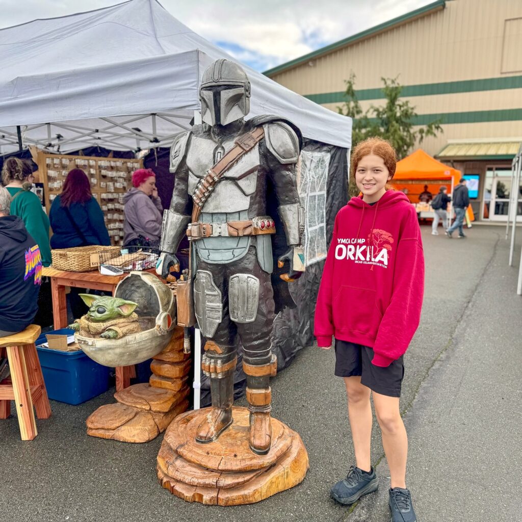 A person in a red hoodie poses next to a wooden sculpture of Star Wars characters at an outdoor art market.