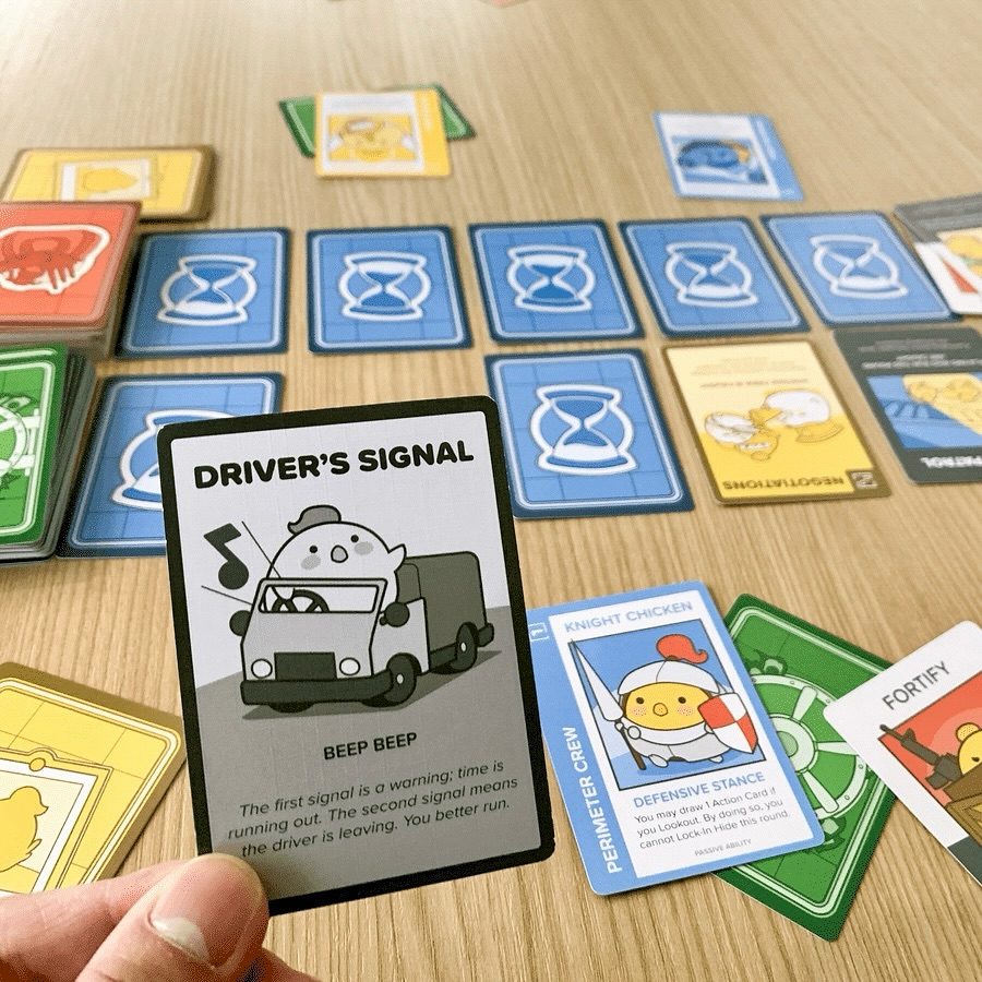 A hand holds a 'Driver's Signal' card from the card game Sumo Penguin, with many colorful game tiles laid out on a light wooden table.