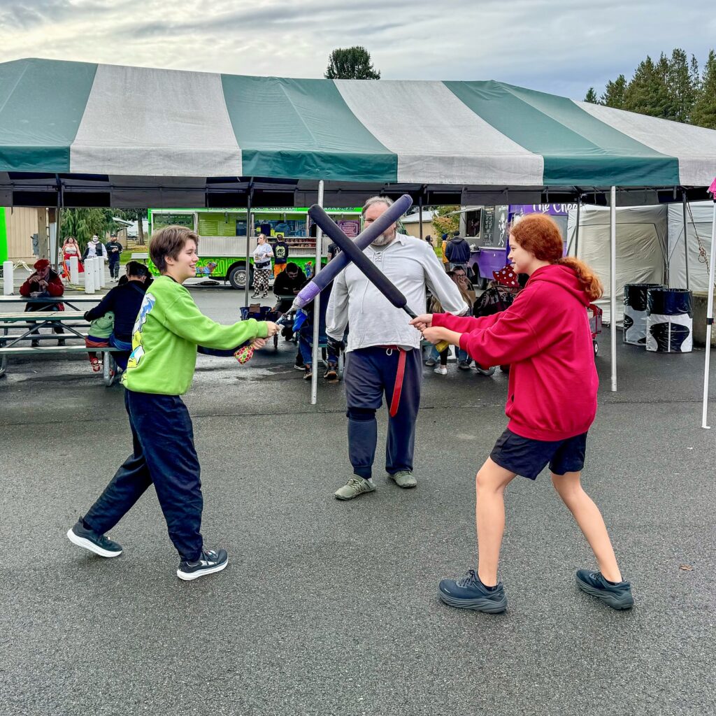 A young boy in a green shirt and a girl in a red hoodie spar with foam swords at an outdoor festival. An instructor watches the practice under a striped canopy.