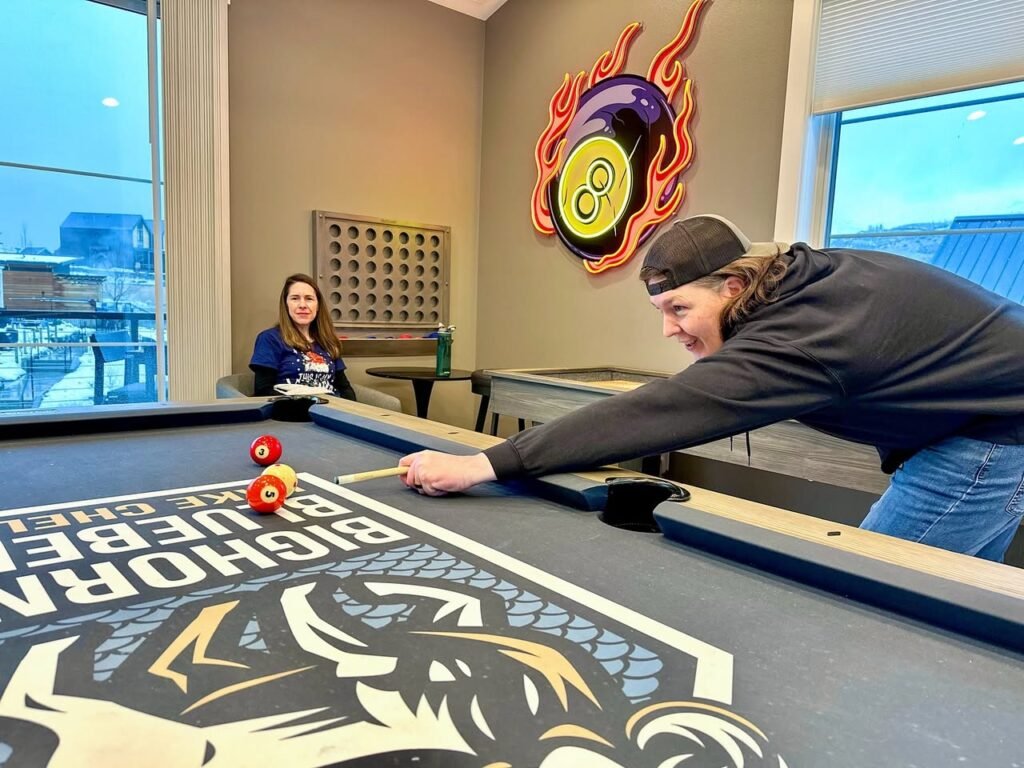 A friend leans over a billiard table with a Buffalo Bills design as another friend watches from across the room.