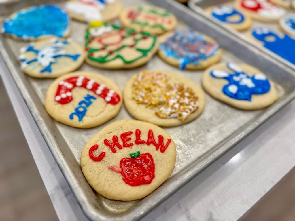 Assorted sugar cookies with festive designs featuring text and holiday symbols on baking sheets.