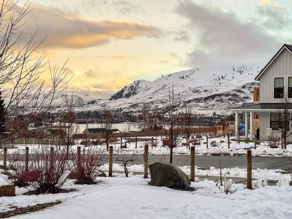 A scenic view of snow-capped mountains and a cozy house in a snowy yard. The setting is picturesque with colorful clouds and a peaceful atmosphere.