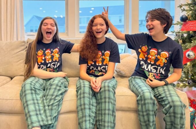 Three kids wearing matching gingerbread Christmas pajamas sit on a couch, laughing and having fun near a decorated holiday tree.