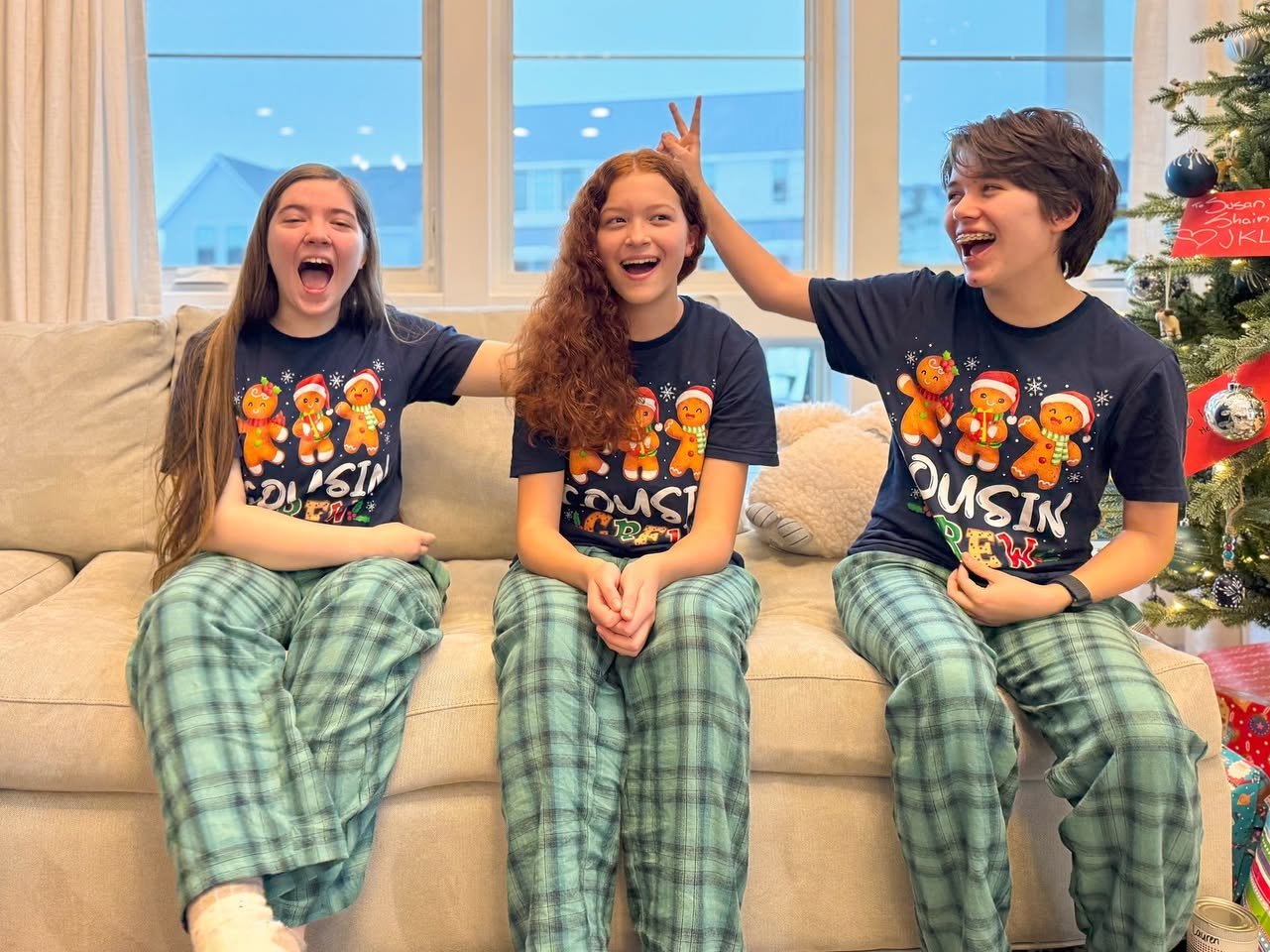 Three kids wearing matching gingerbread Christmas pajamas sit on a couch, laughing and having fun near a decorated holiday tree.