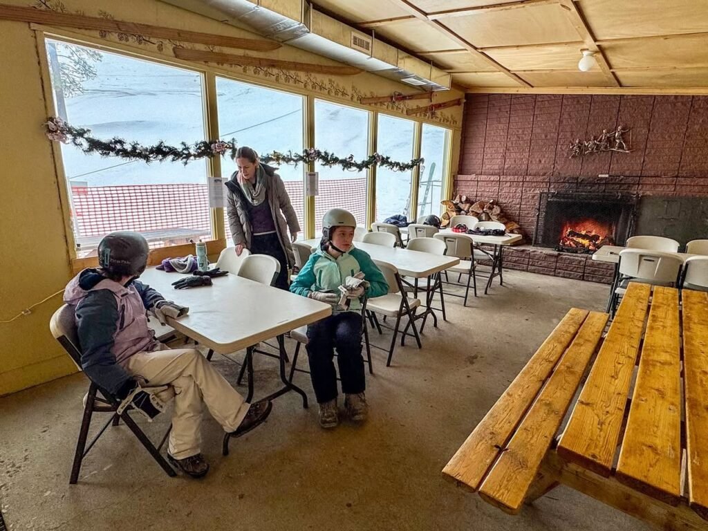 People gathered indoors near a fireplace in a rustic lodge with large windows.