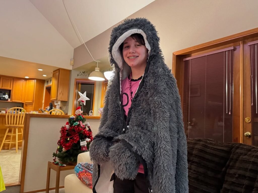 A person wearing a fluffy gray hooded coat stands in a warmly lit living room with holiday decorations.