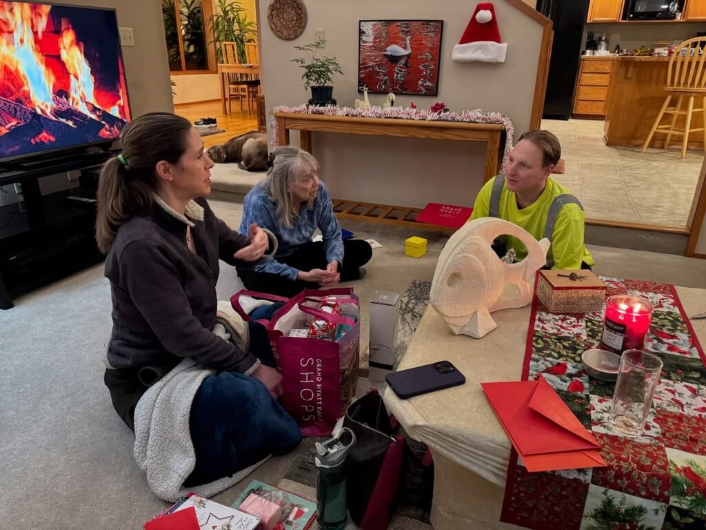 A group of relatives gathers on the living room floor to open presents during the holidays. A TV displays a cozy fireplace alongside festive decorations and holiday gifts.