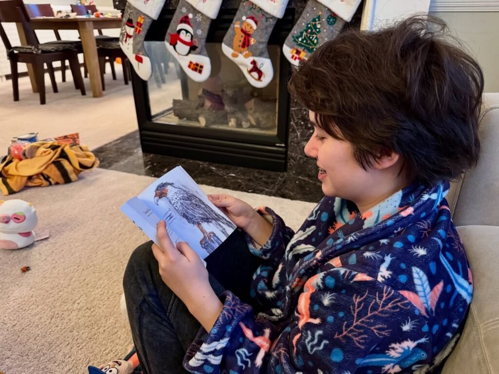 A person wearing a festive flannel shirt reads a card by a fireplace adorned with Christmas stockings, capturing a cozy holiday moment.