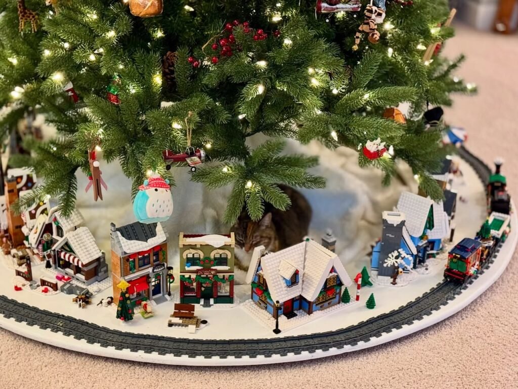 A miniature snow-covered village with tiny houses and a toy train loop surrounds the base of a decorated Christmas tree. Close-up details highlight festive miniature structures and twinkling lights.