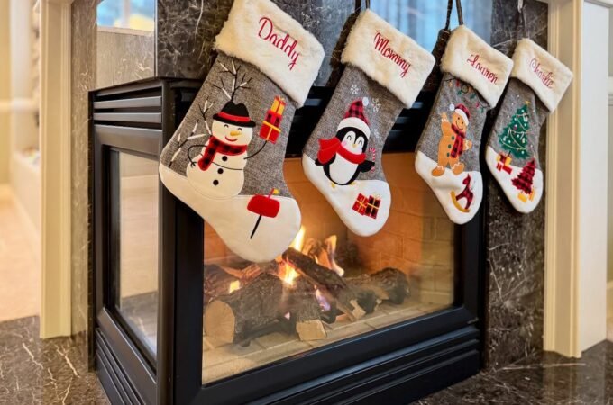 Four Christmas stockings with snowman and penguin designs hang above a modern fireplace.