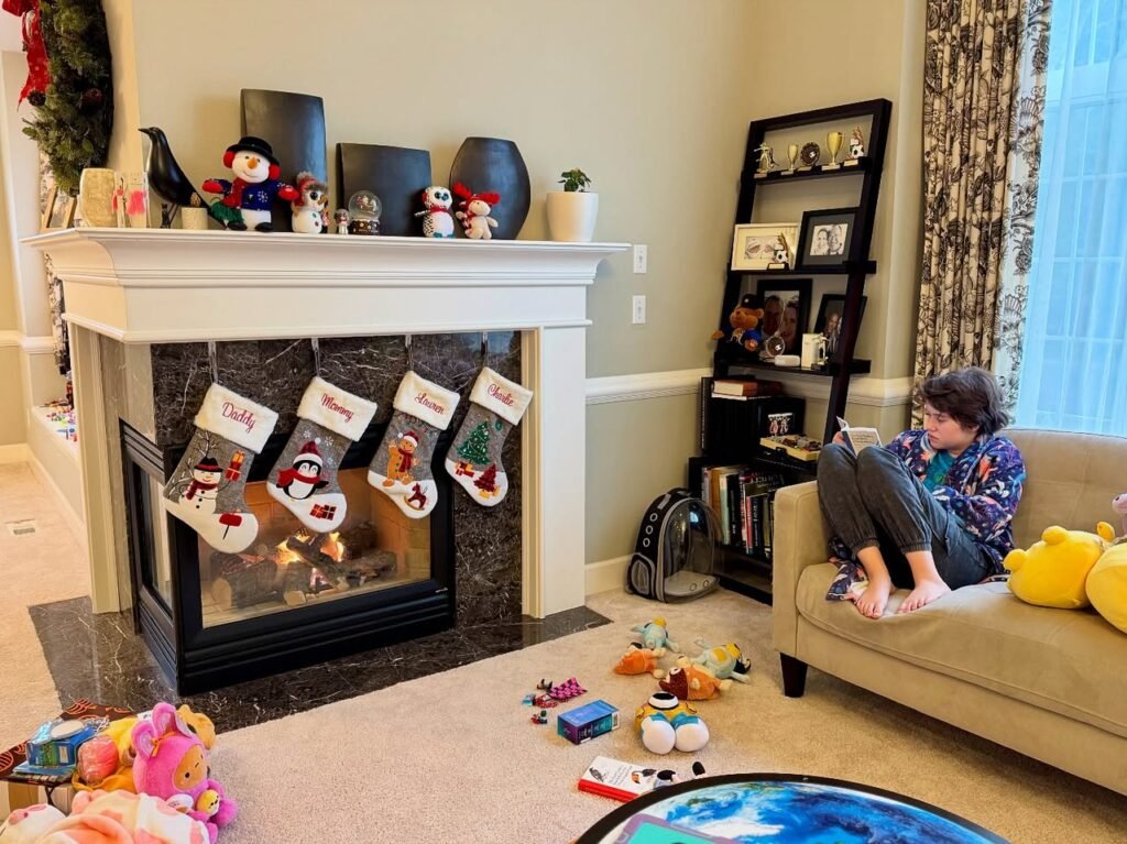 A cozy living room features a white fireplace mantel adorned with snowman figurines, stockings, and holiday decorations. A person relaxes on a beige sofa nearby.