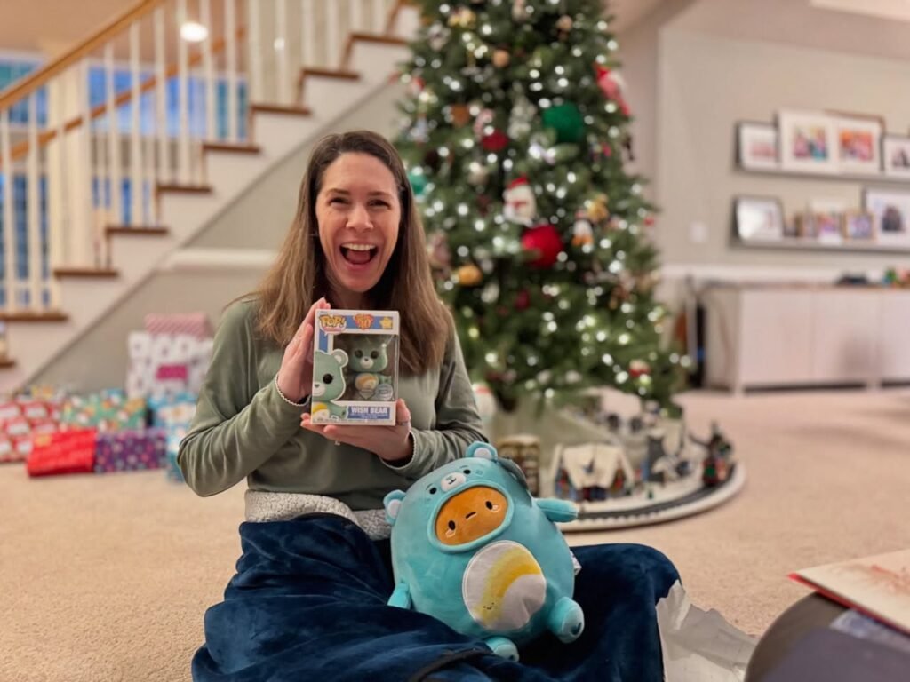 A smiling person celebrating the arrival of Christmas morning by holding a small gift box and a large plush toy in a cozy living room.
