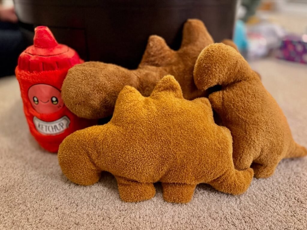 Red and brown plush toys sitting together on a carpet. A red bottle-shaped plush with a face is next to brown dinosaur plushes.