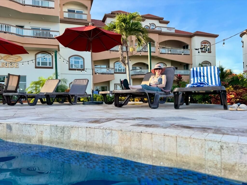 A person lounges on a sunbed by a swimming pool, shaded by red umbrellas under a clear blue sky.