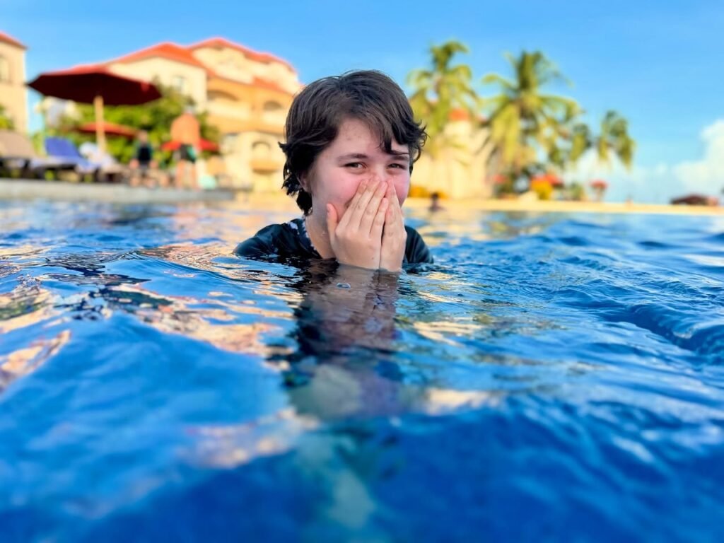 Children swimming in a tropical resort pool, covering their mouths in laughter