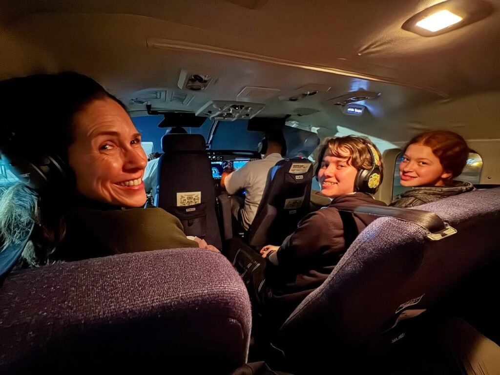 A group of smiling people wearing headsets, looking out the window of an airplane cabin at night.