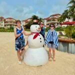 A group of three people poses with a snowman prop on a sandy beach near a resort with palm trees.