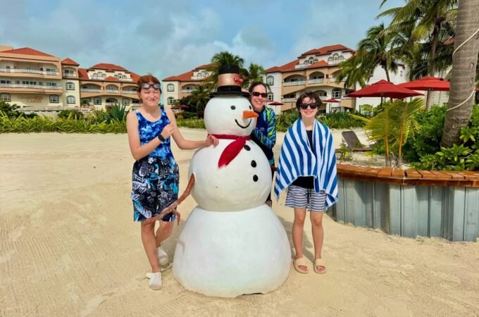 A group of three people poses with a snowman prop on a sandy beach near a resort with palm trees.