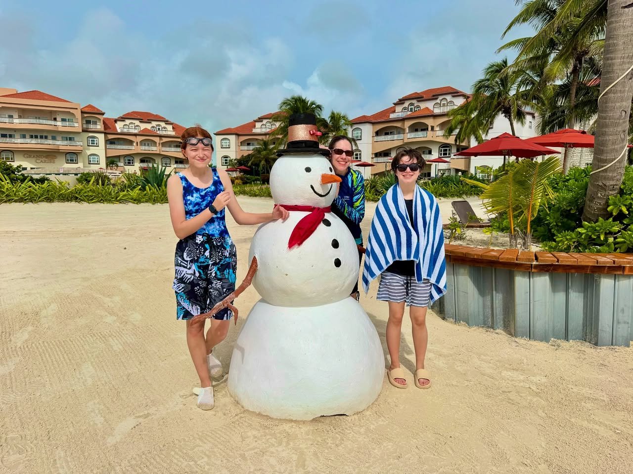 A group of three people poses with a snowman prop on a sandy beach near a resort with palm trees.
