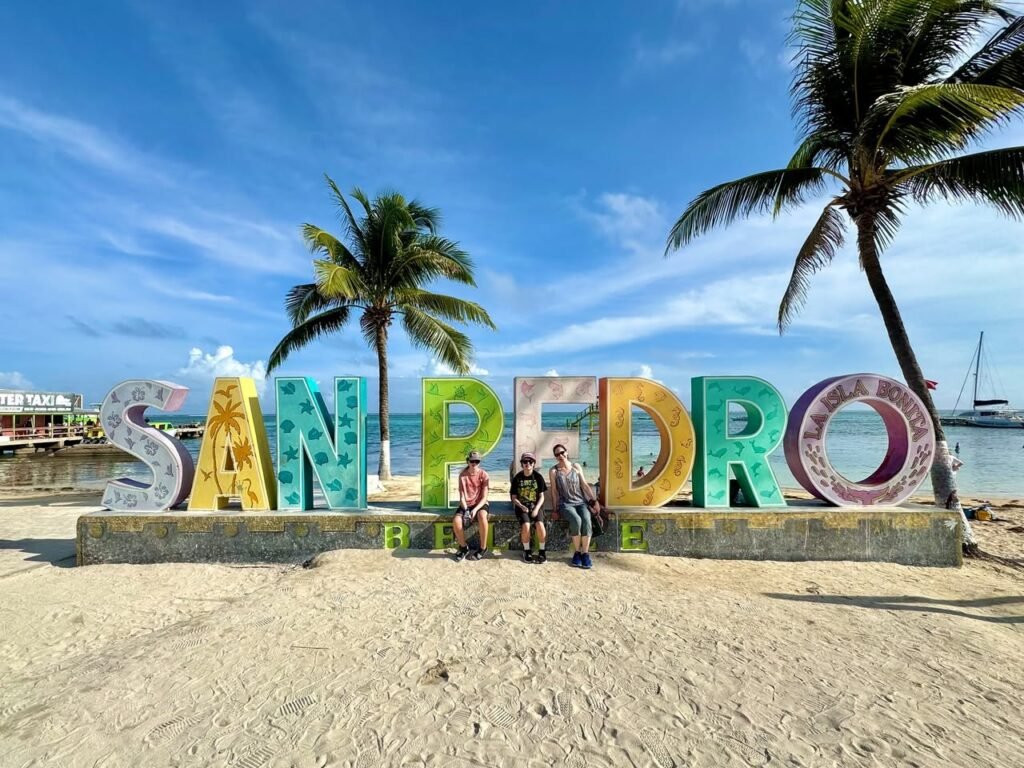 A family poses in front of a colorful San Pedro sign on a sandy beach. Palm trees frame the photo under a clear blue sky.