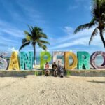A family poses in front of a colorful San Pedro sign on a sandy beach. Palm trees frame the photo under a clear blue sky.