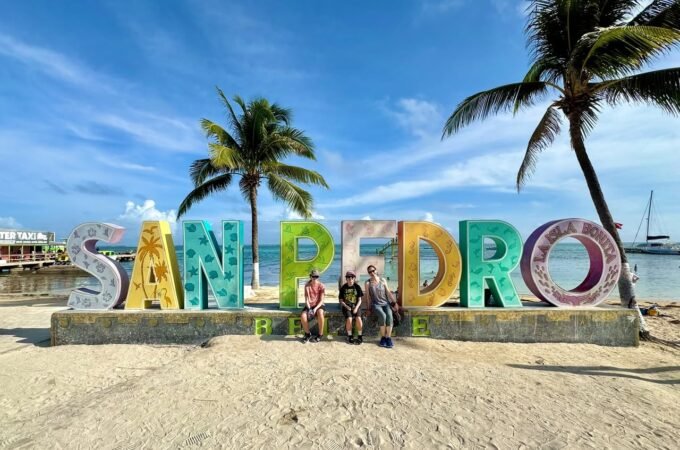 A family poses in front of a colorful San Pedro sign on a sandy beach. Palm trees frame the photo under a clear blue sky.