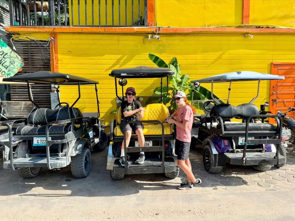 Two people pose with a golf cart next to a bright yellow building.