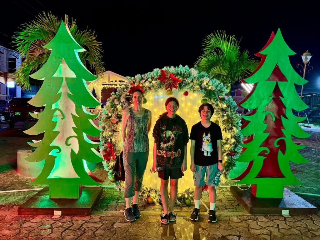 Three people posing for a holiday photo next to large illuminated Christmas tree decorations and a lighted arch at night.