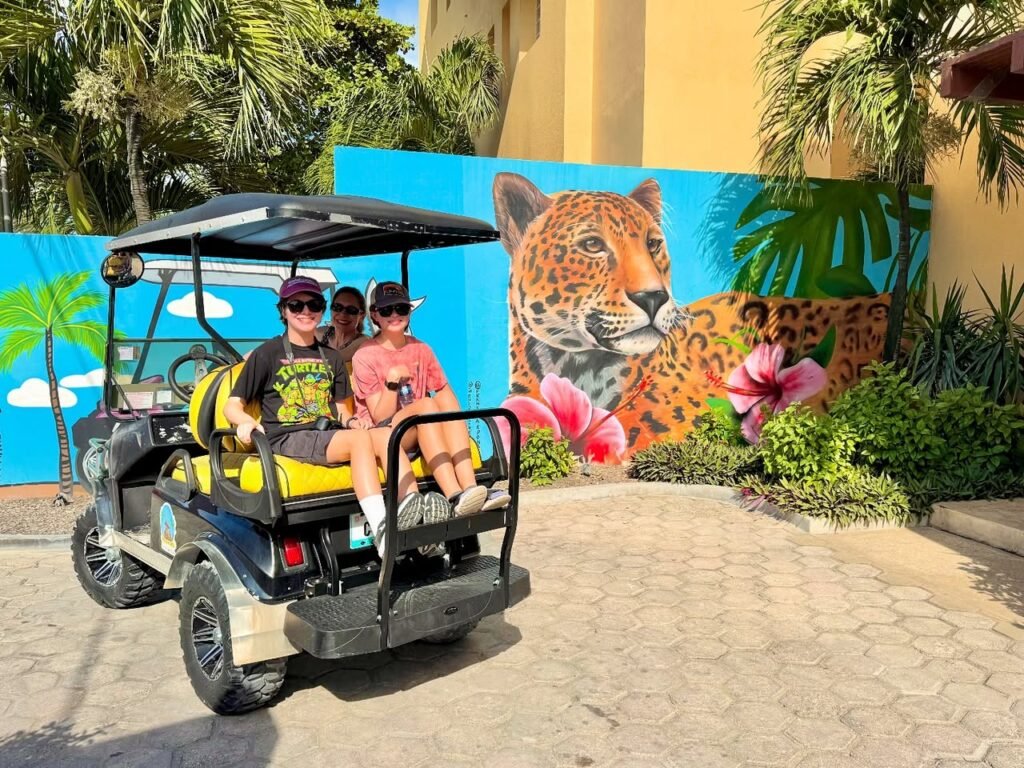 A group rides a golf cart along a paved path near a vibrant leopard mural with tropical foliage.