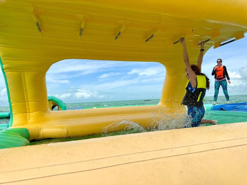 Two people wearing life jackets on a large yellow and green inflatable water park by a lake.