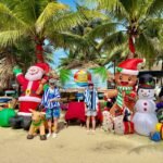 A family enjoys a beachside Christmas photo op in Puerto Vallarta, Mexico, surrounded by festive inflatable holiday decorations under palm trees.