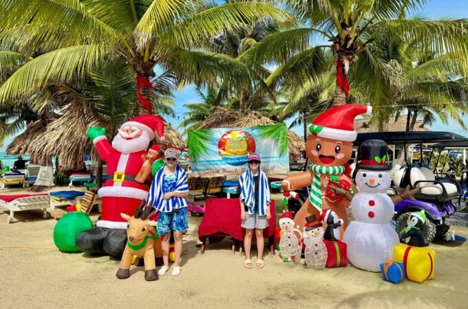 A family enjoys a beachside Christmas photo op in Puerto Vallarta, Mexico, surrounded by festive inflatable holiday decorations under palm trees.