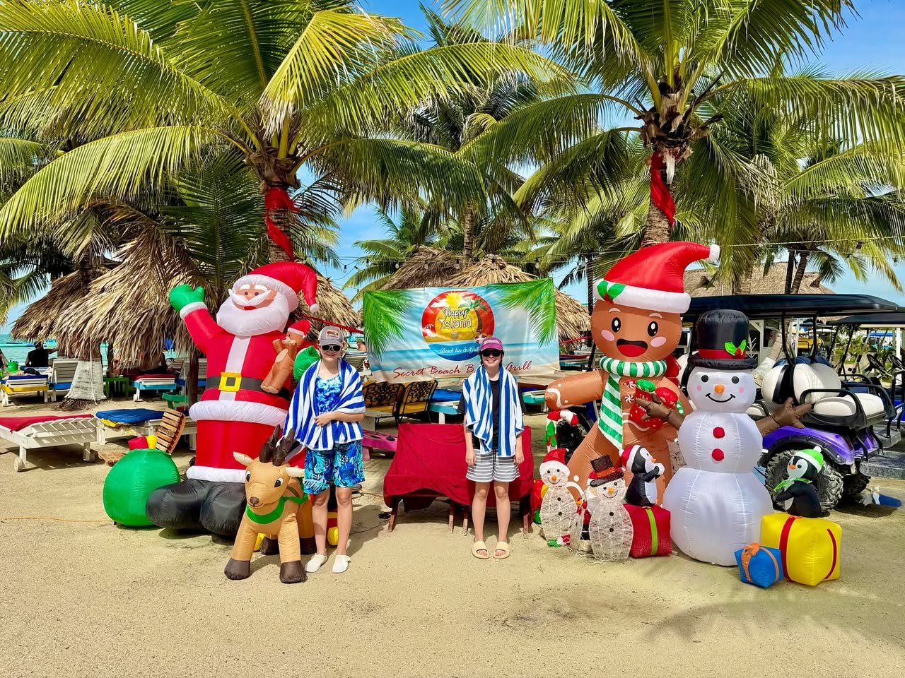 A family enjoys a beachside Christmas photo op in Puerto Vallarta, Mexico, surrounded by festive inflatable holiday decorations under palm trees.