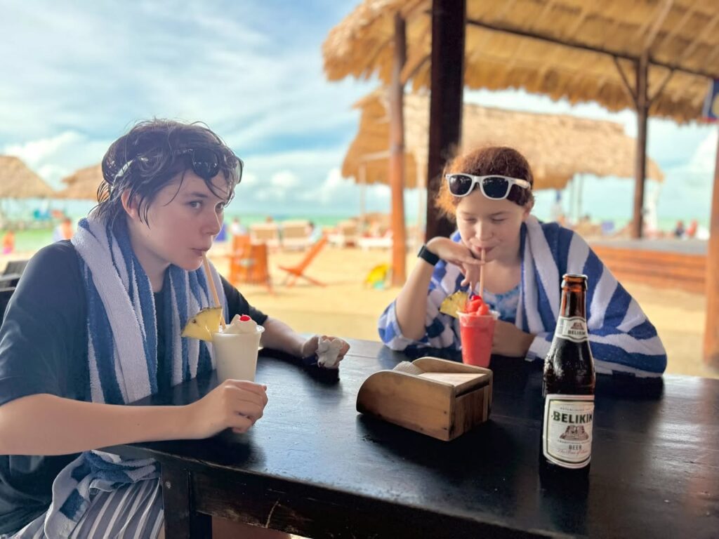 A couple enjoys cocktails and beer at an outdoor tropical bar with thatched umbrellas and ocean views.