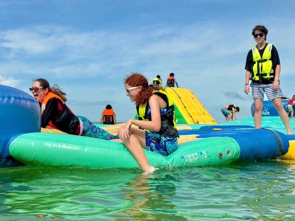 A group of people enjoy an inflatable water obstacle course in a lake, wearing life jackets under a bright blue sky.
