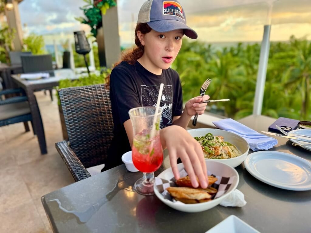 A person enjoys a meal with tropical drinks and potato wedges on an open-air patio. The lush green surroundings enhance the relaxed dining experience.